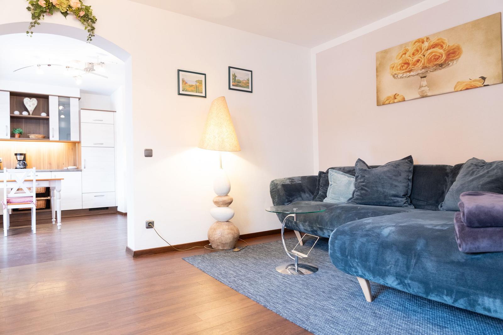 Wohnzimmer mit Sofa, Beistelltisch und Stehlampe. Küche im Hintergrund sichtbar.