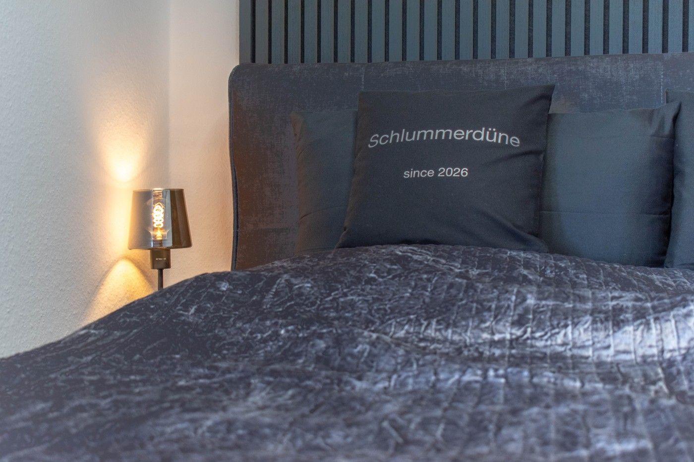 Bettkopfteil mit Kissen, auf dem Schlummerdüne since 2026 steht, und Nachttischlampe.