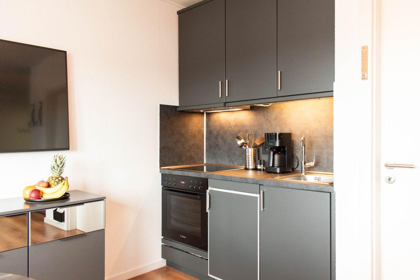 Küche mit dunklen Schränken, Herd und Spüle. Fernseher und Obst auf Sideboard.