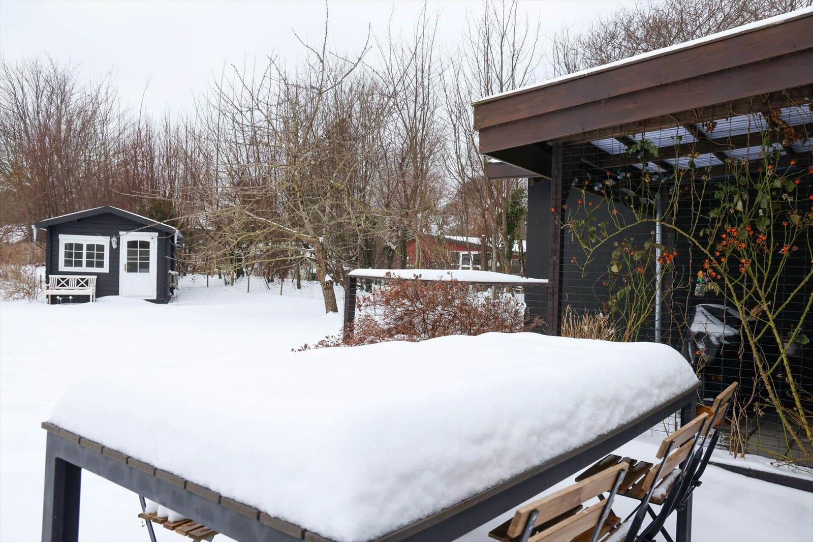 Schnee bedeckt einen Tisch und Stühle vor einem Haus mit schwarzem Dach und einem Garten.