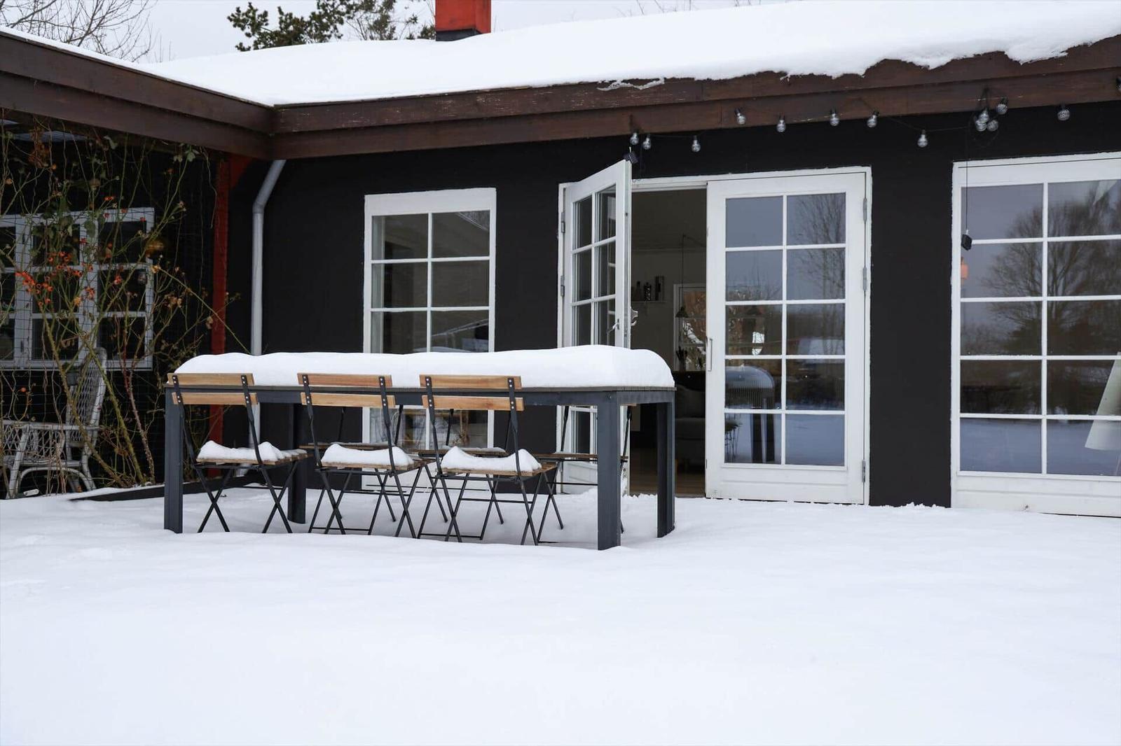 Schneebedeckter Außenbereich mit Tisch und Stühlen vor einem Haus mit weißen Fenstern.