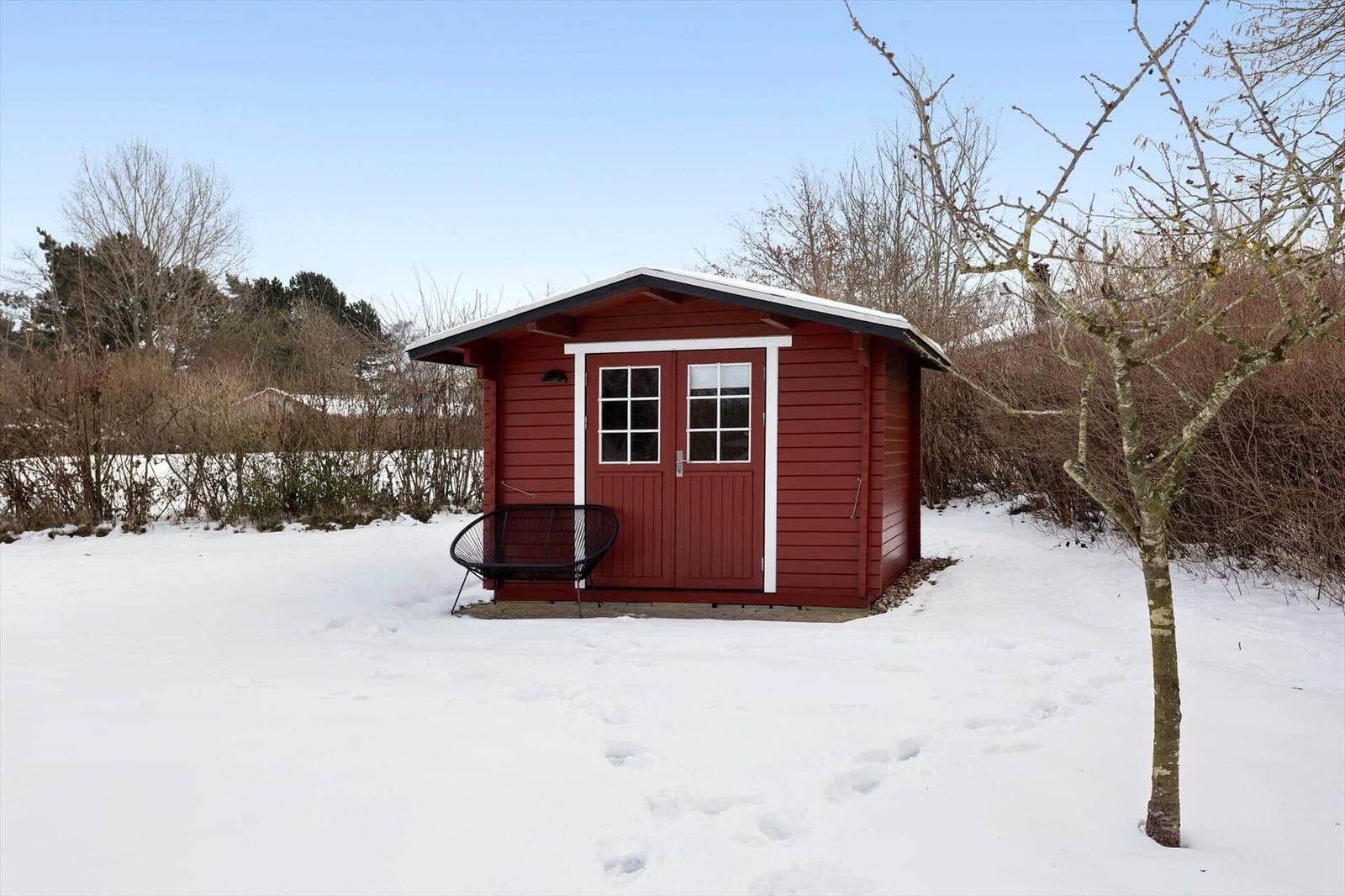 Red wooden house with snow-covered ground and a black chair in front of the door.