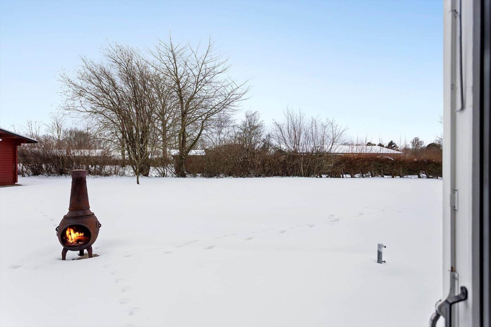 Snow-covered garden with a lit outdoor fireplace and bare trees in the background.