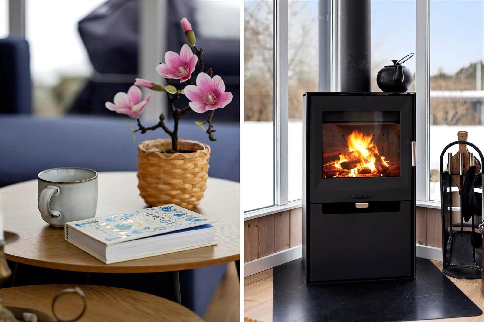 Table with flower, mug, and book. Fireplace with burning wood and window.