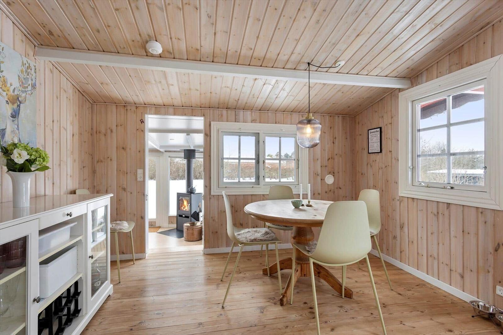 Dining room with wooden walls, round table, chairs, windows, and view into living area.