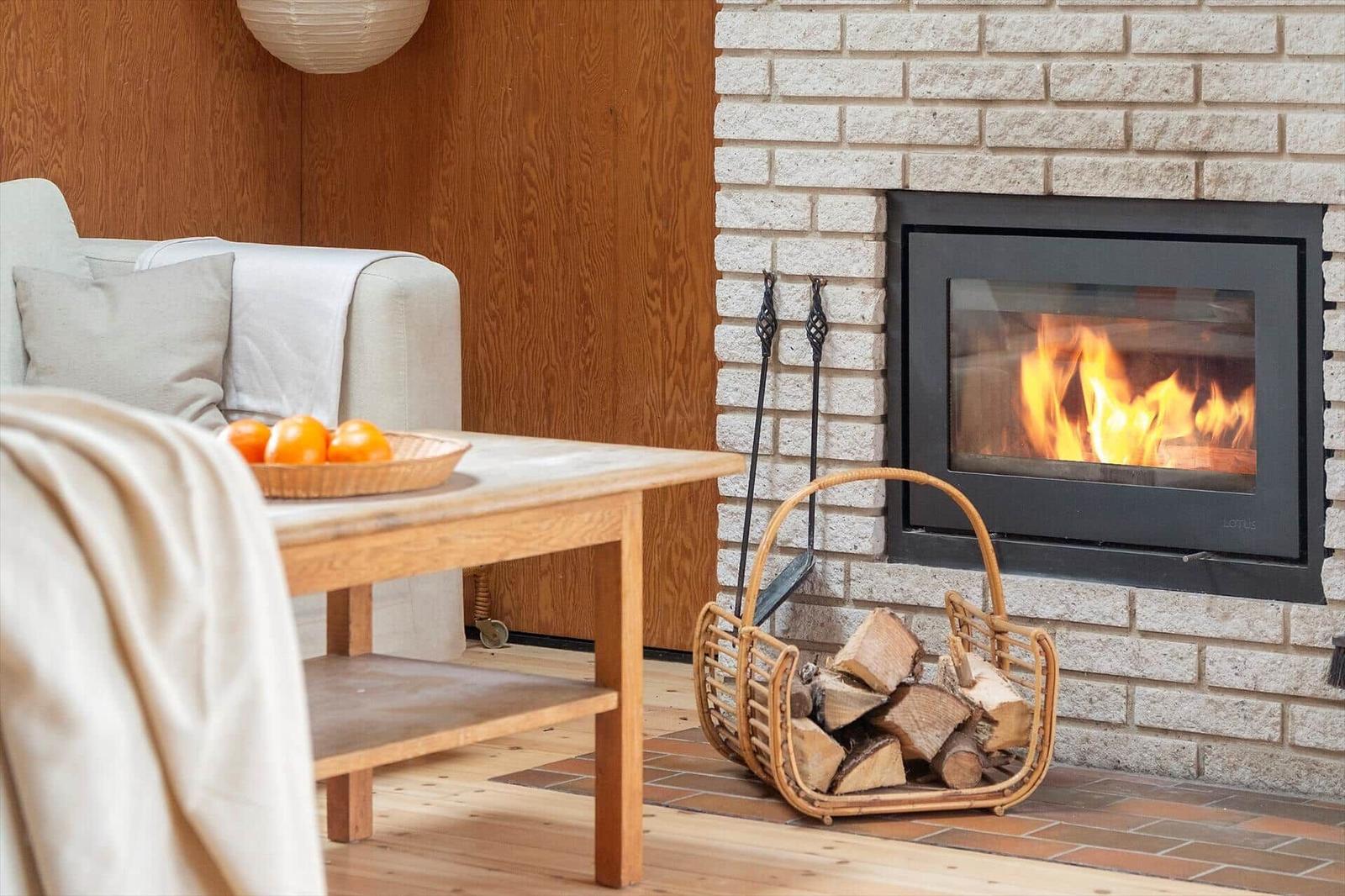 A fireplace with burning fire, wood in a basket, and a sofa with cushions.