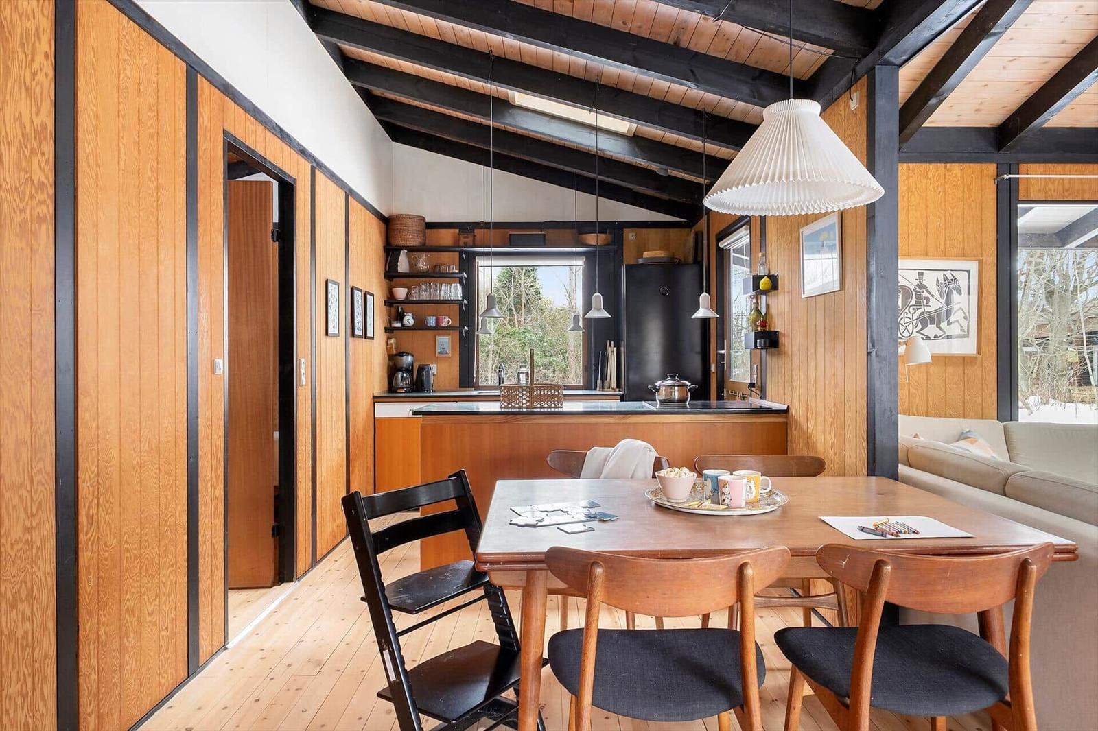 Dining area with wooden table and chairs, kitchen in background, wood walls and floor.