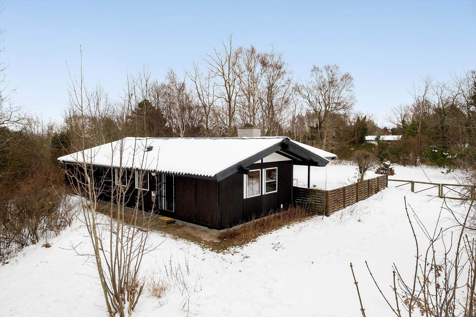 Black wooden house with snow-covered roof and garden fence.