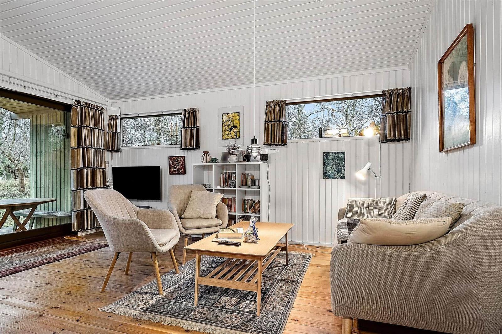 Wohnzimmer mit Sofa, Sesseln, Couchtisch und Bücherregal. Holzboden und Fenster mit Vorhängen.