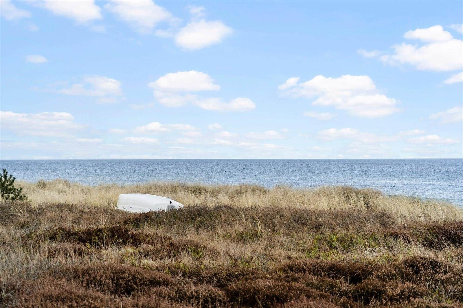 Ein weißes Boot liegt im hohen Gras am Strand. Der Horizont ist weit und der Himmel blau mit weißen Wolken.