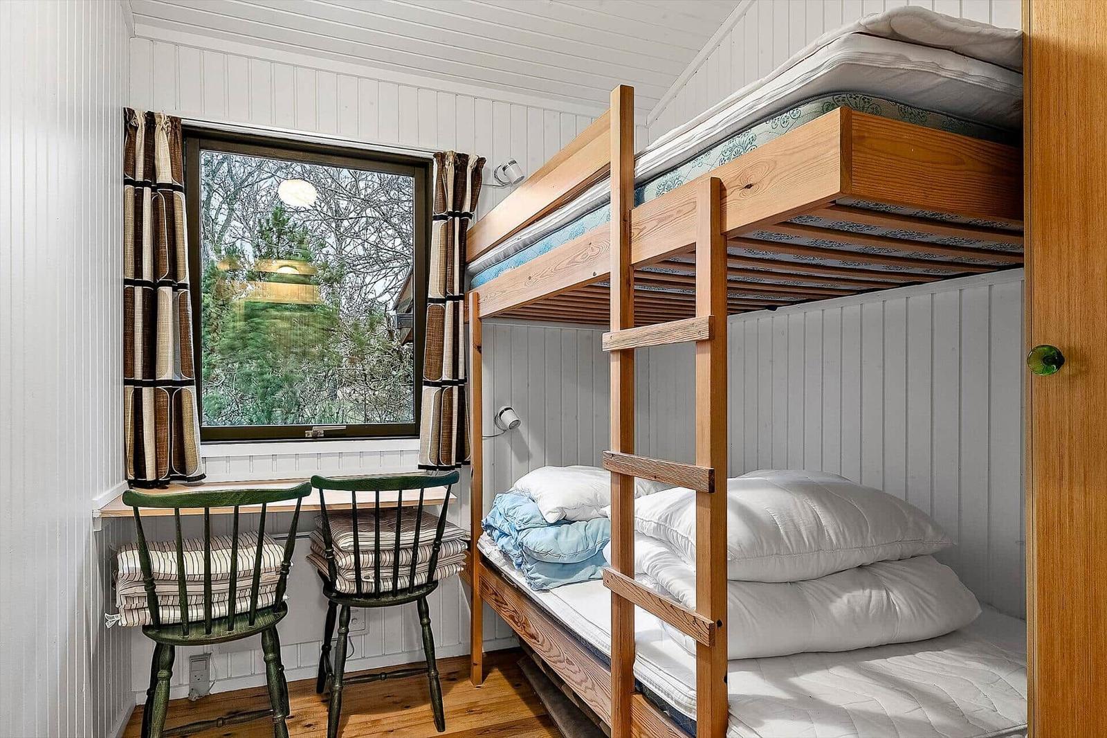 Zimmer mit Schlafcouch, Tisch und zwei Stühlen. Fenster mit Vorhängen. Holzboden.