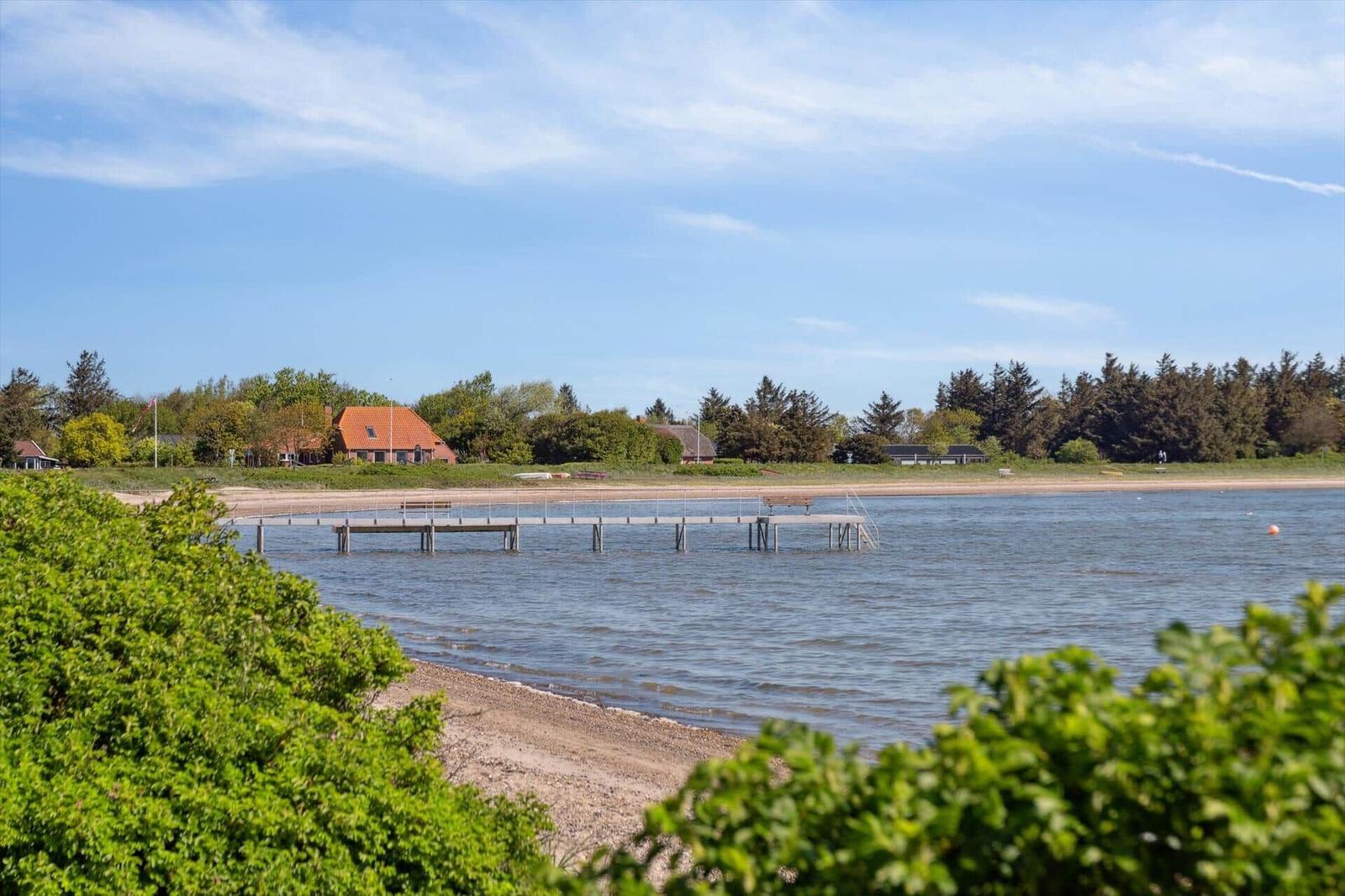 Hafen mit Steg, Strand und Häusern im Hintergrund unter blauem Himmel.