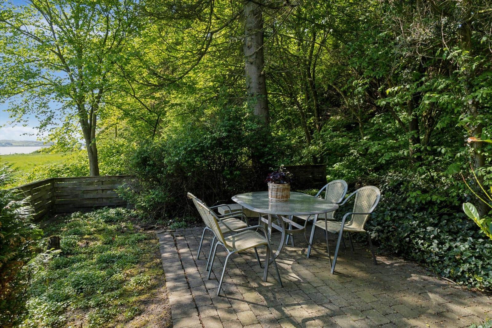 Garten mit Tisch und Stühlen, umgeben von Bäumen und Grünflächen. Blick auf Wasser im Hintergrund.