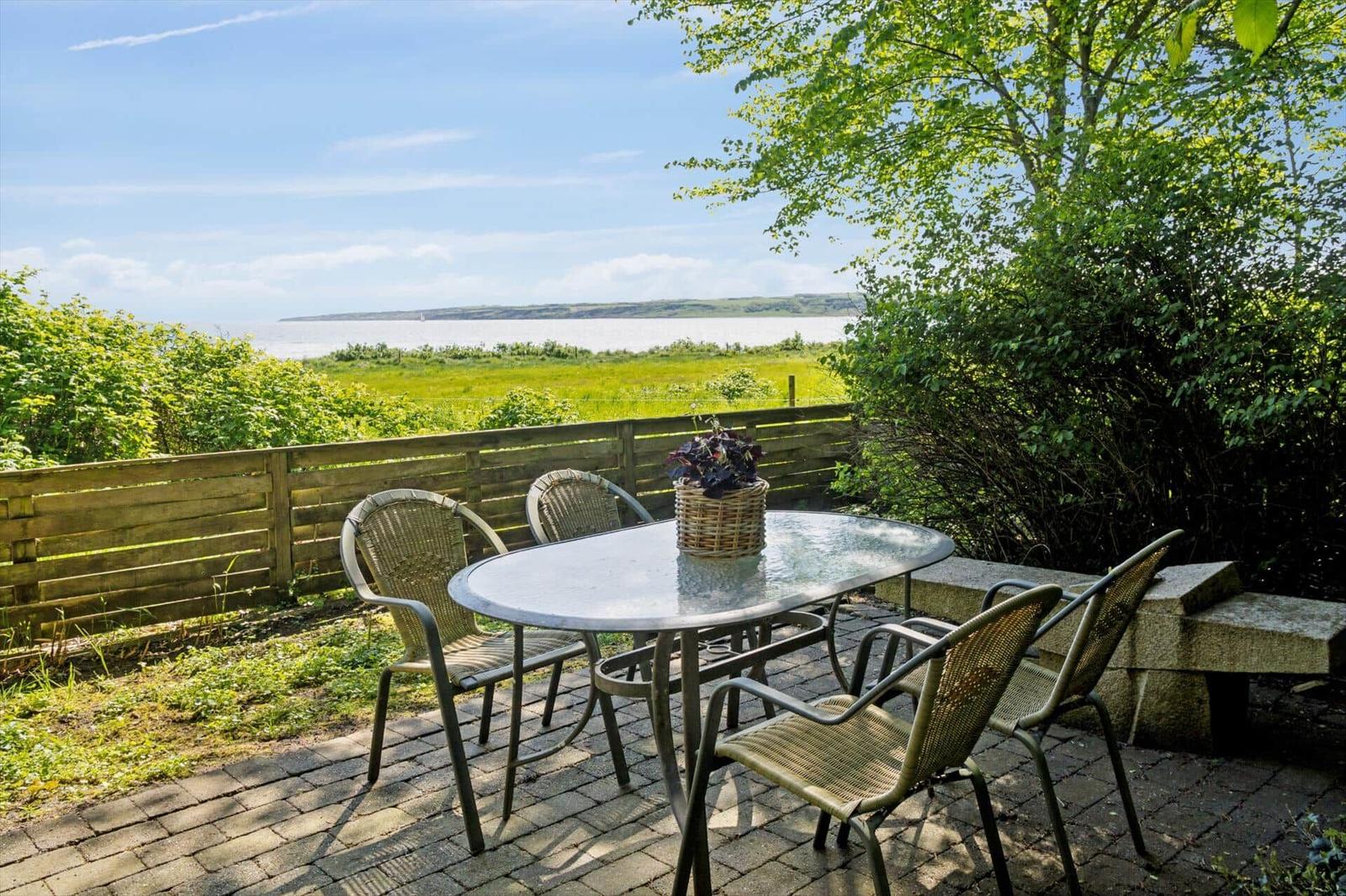 Terrasse mit Tisch und Stühlen, Blick auf Wasser und Grünflächen.
