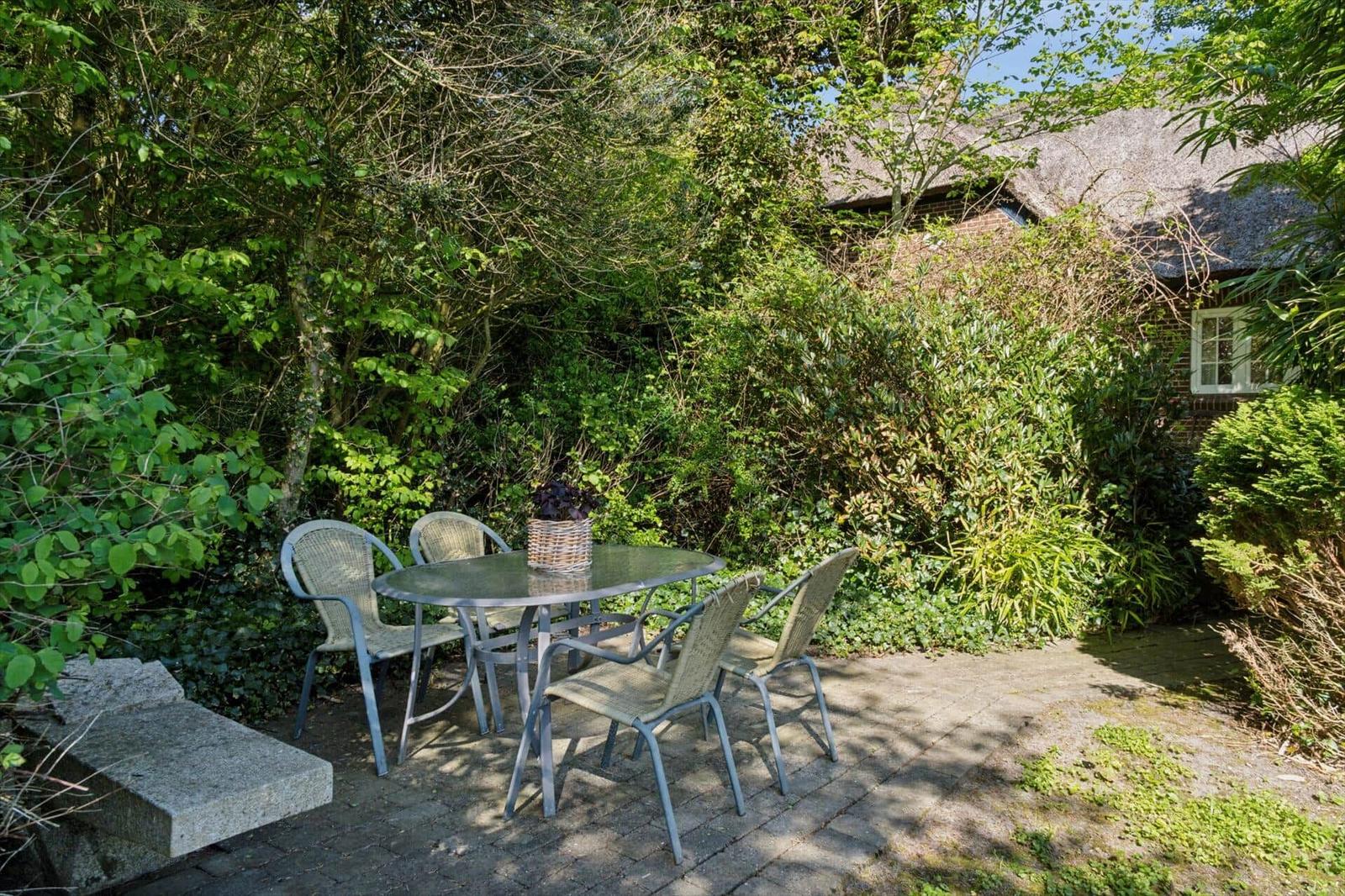Terrasse mit Tisch und Stühlen, umgeben von grüner Vegetation und einem Haus mit Schilfdach.