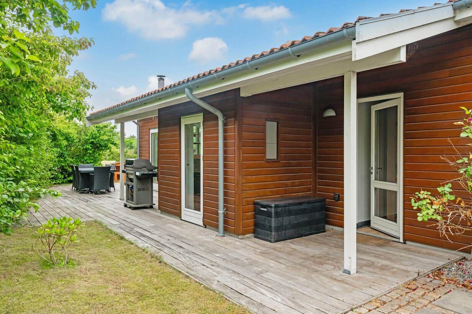 Wooden house with terrace, grill, and garden furniture under blue sky.