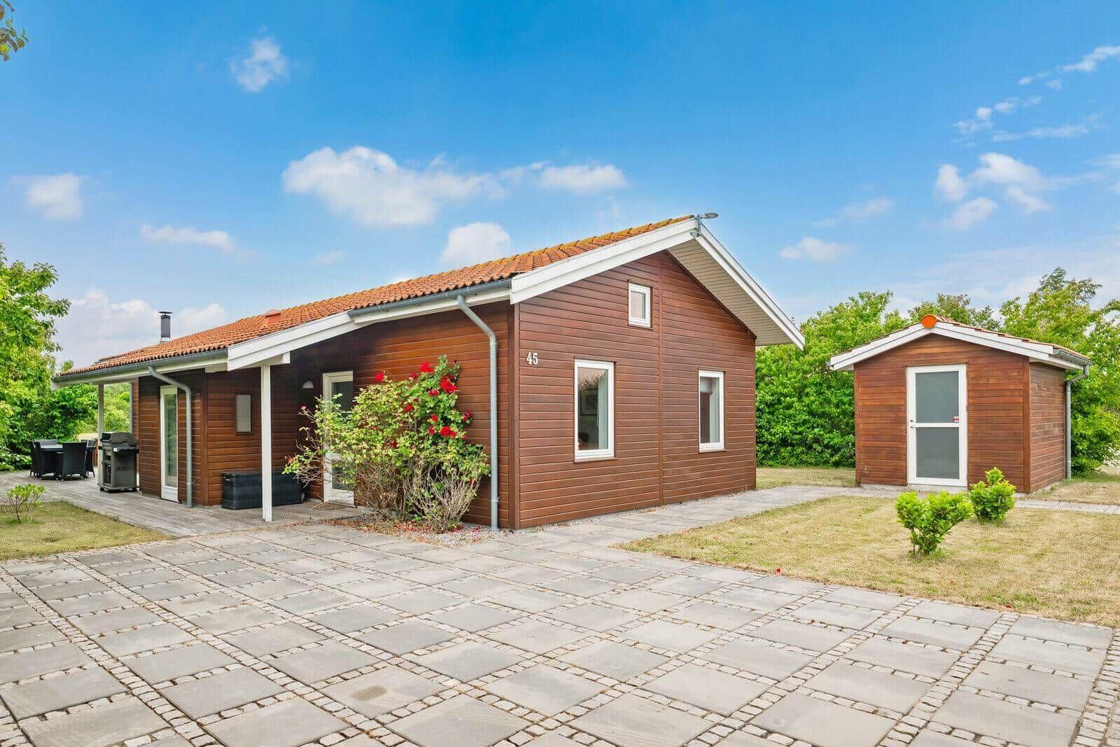 Wooden house with terrace, grill, and small outbuilding on paved grounds.