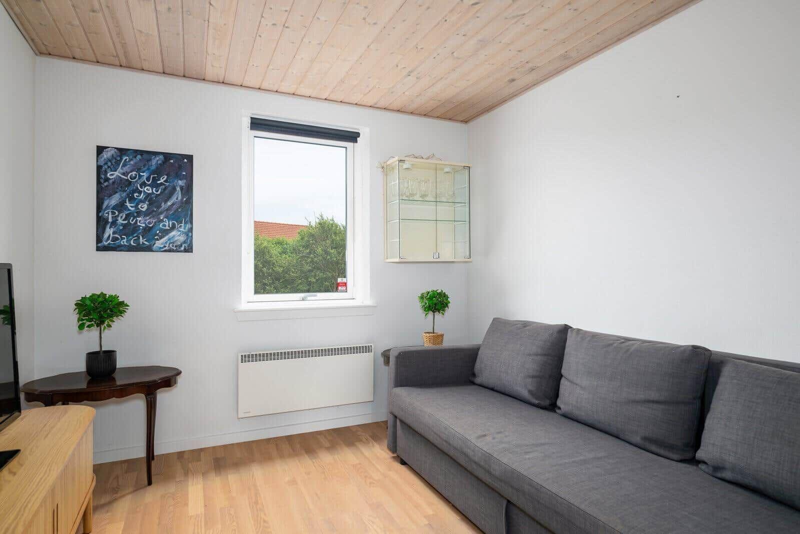 Living room with gray sofa, wooden floor, and wooden ceiling. Window with view of greenery.