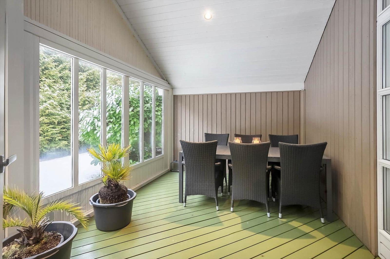 Terrace with table, six chairs, and plants. Large window front with view of greenery.