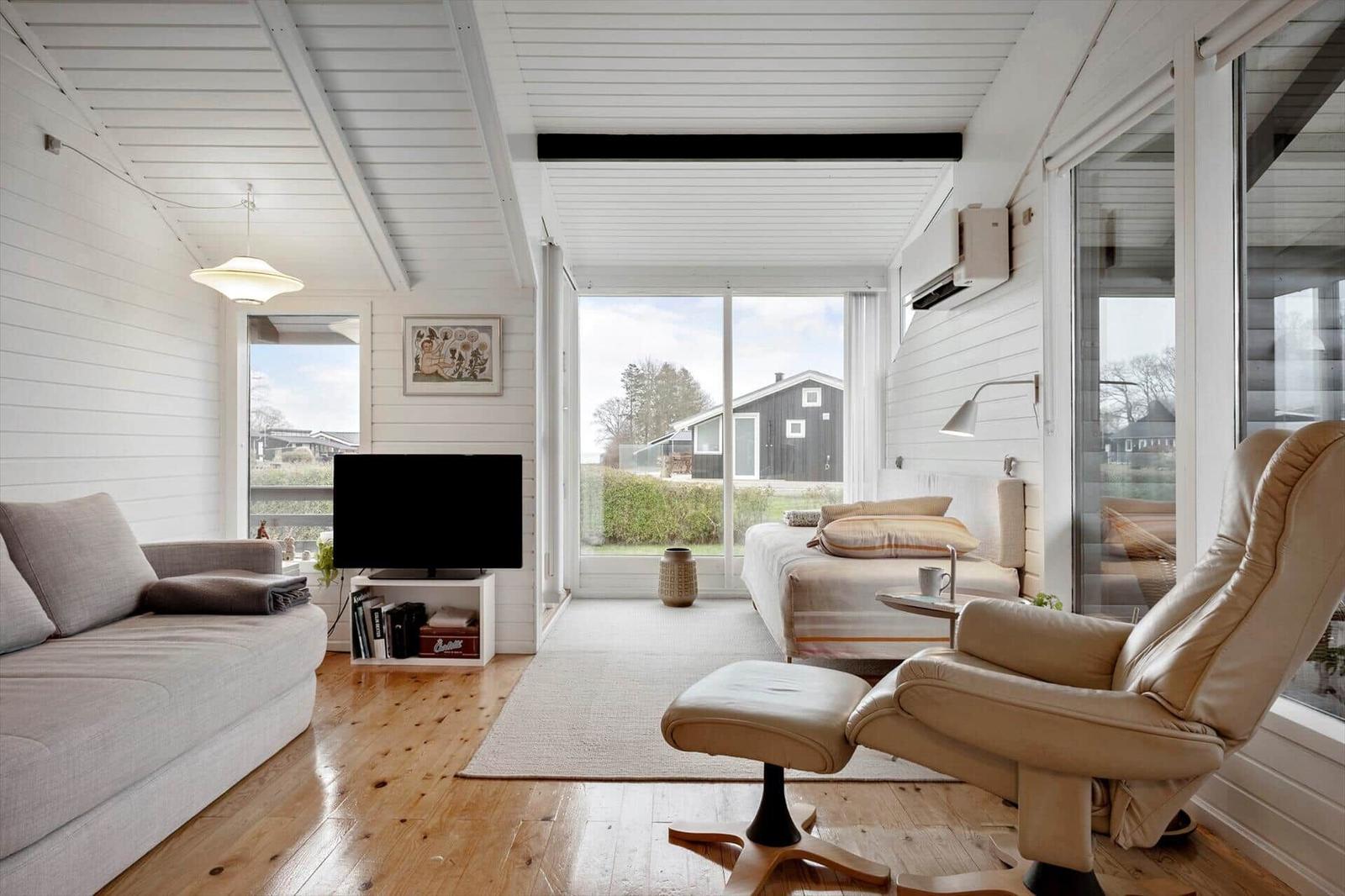 Wohnraum mit Sofa, Fernseher und Sessel. Holzboden und große Fenster.