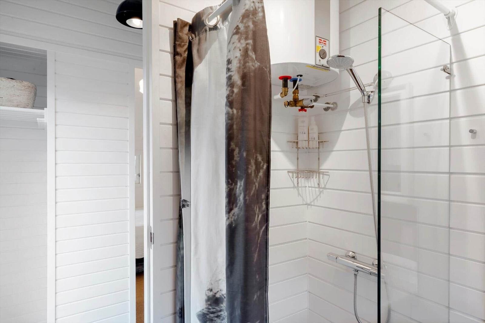 Dusche mit Glaswand, Duschvorhang und Regal. Weiße Fliesen und Heizung.