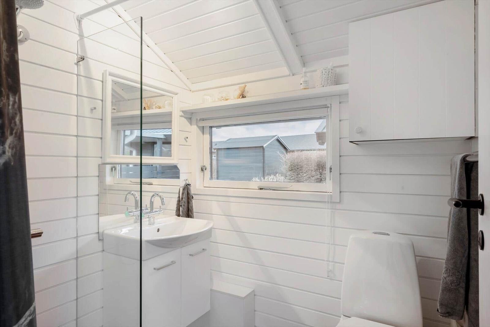 Badezimmer mit Dusche, Waschbecken und Toilette. Weiße Wände und Fenster mit Blick.