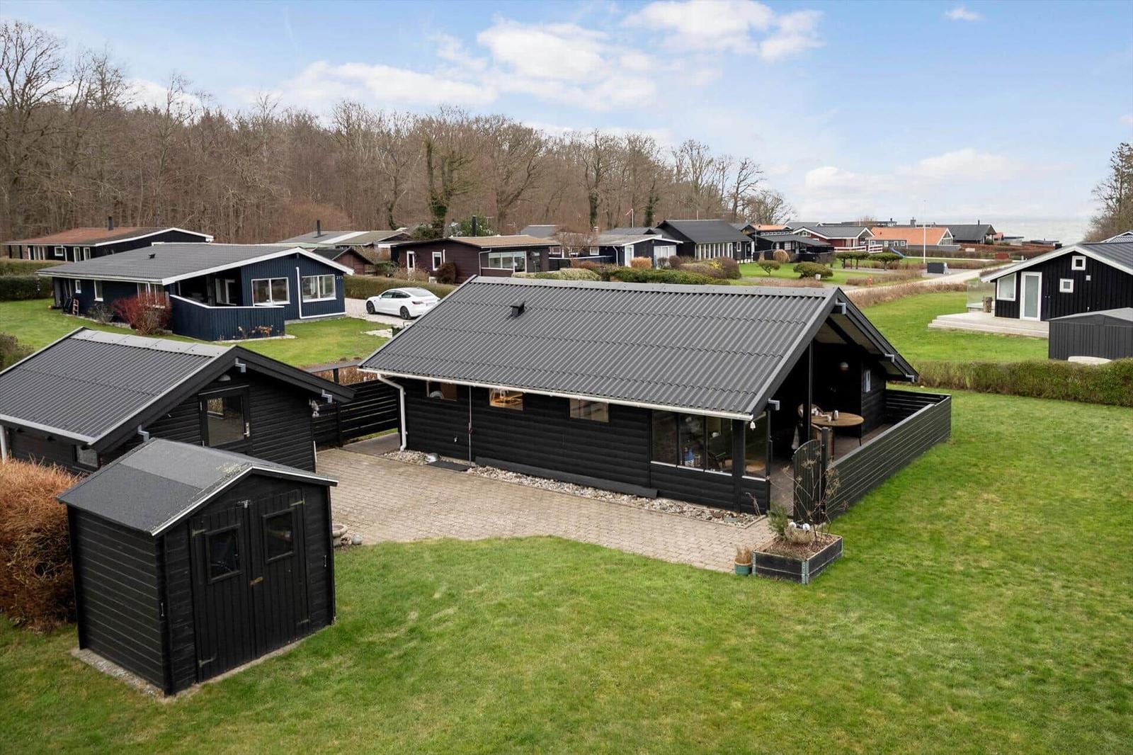 Schwarze Holzhütte mit Terrasse und Garten. Umgebung mit weiteren Häusern und Wald.