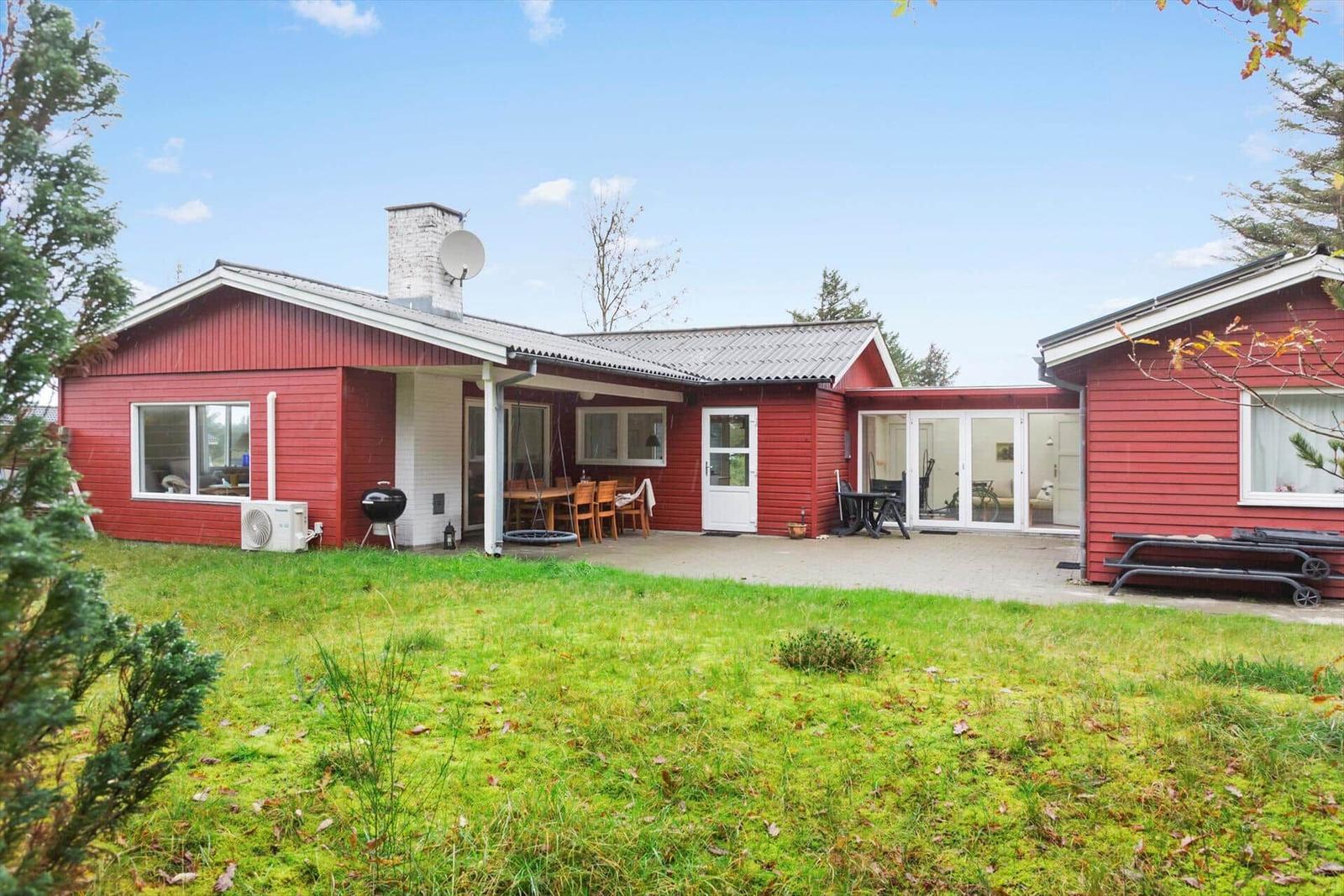 Rotes Ferienhaus mit Terrasse, Grill und Garten. Glasfront öffnet sich zum Innenraum.