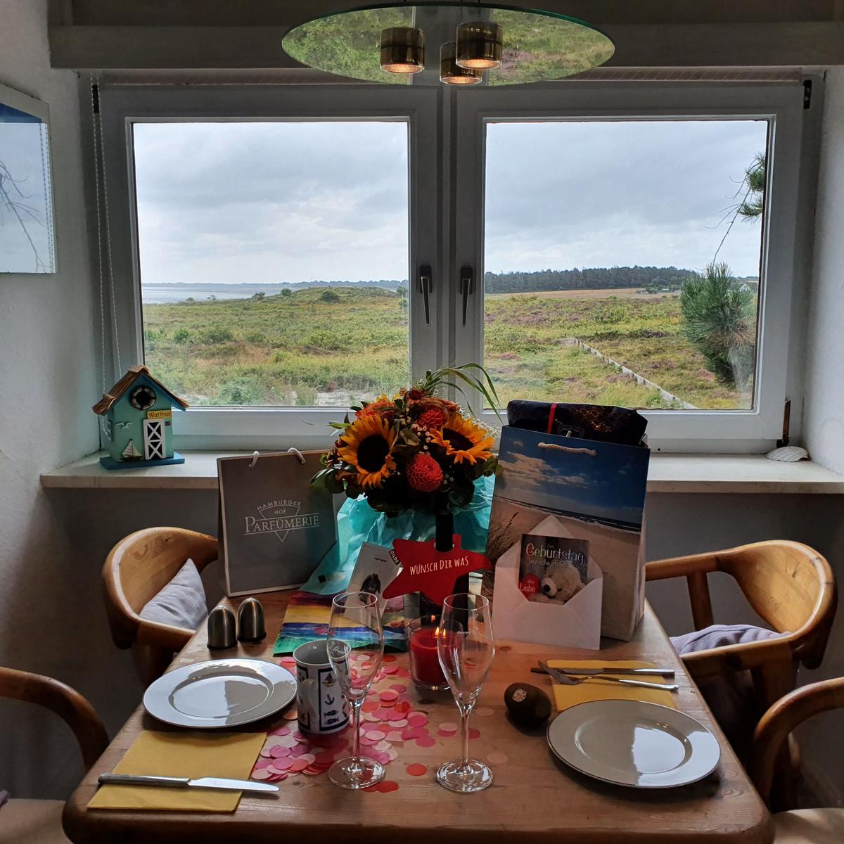 Tisch mit Blumen und Geschenken vor Fenster mit Landschaftsaussicht.