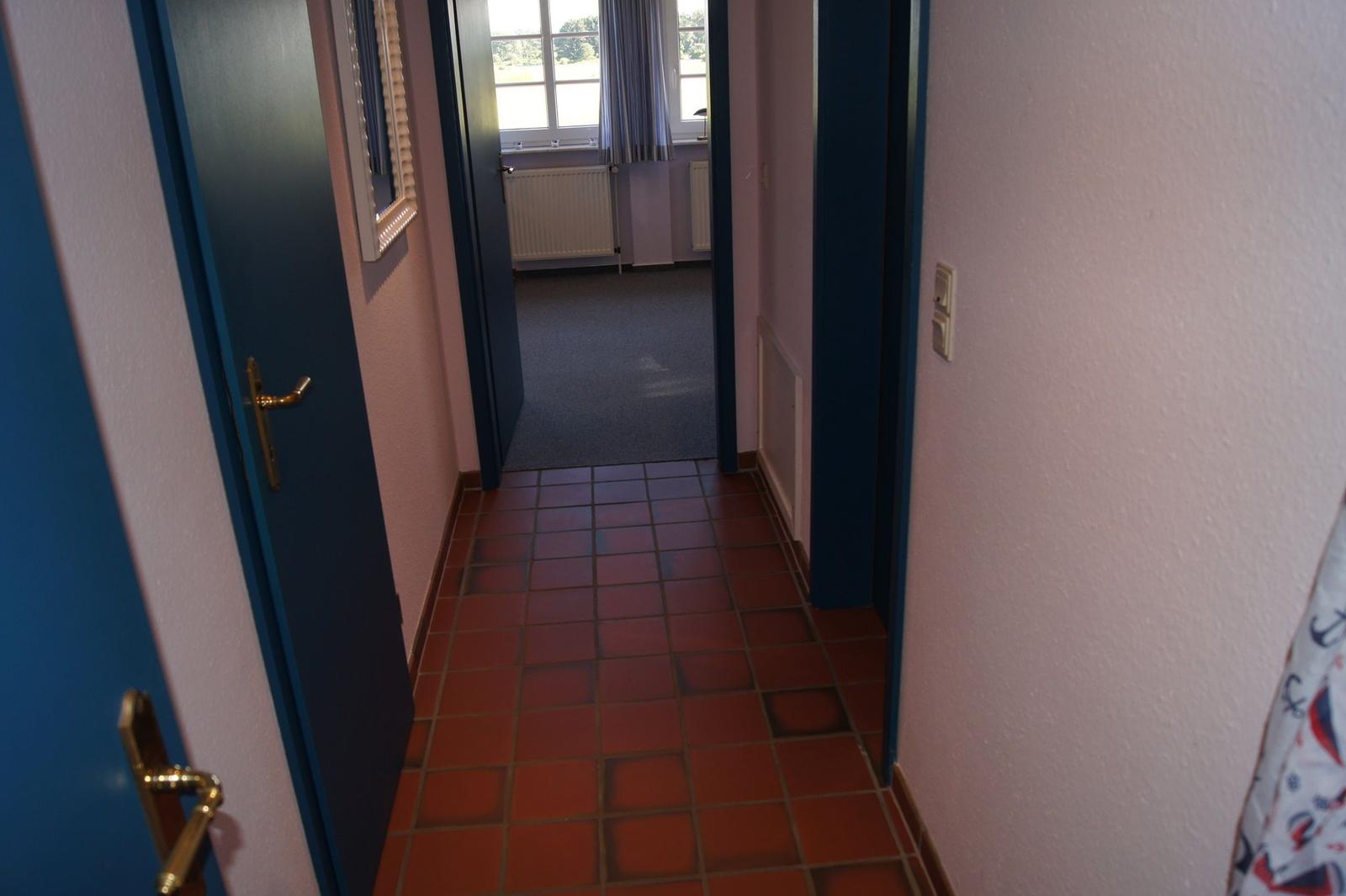 Flur mit roten Fliesen und blauen Türen. Im Hintergrund ist ein Raum mit Fenster und Heizkörper.