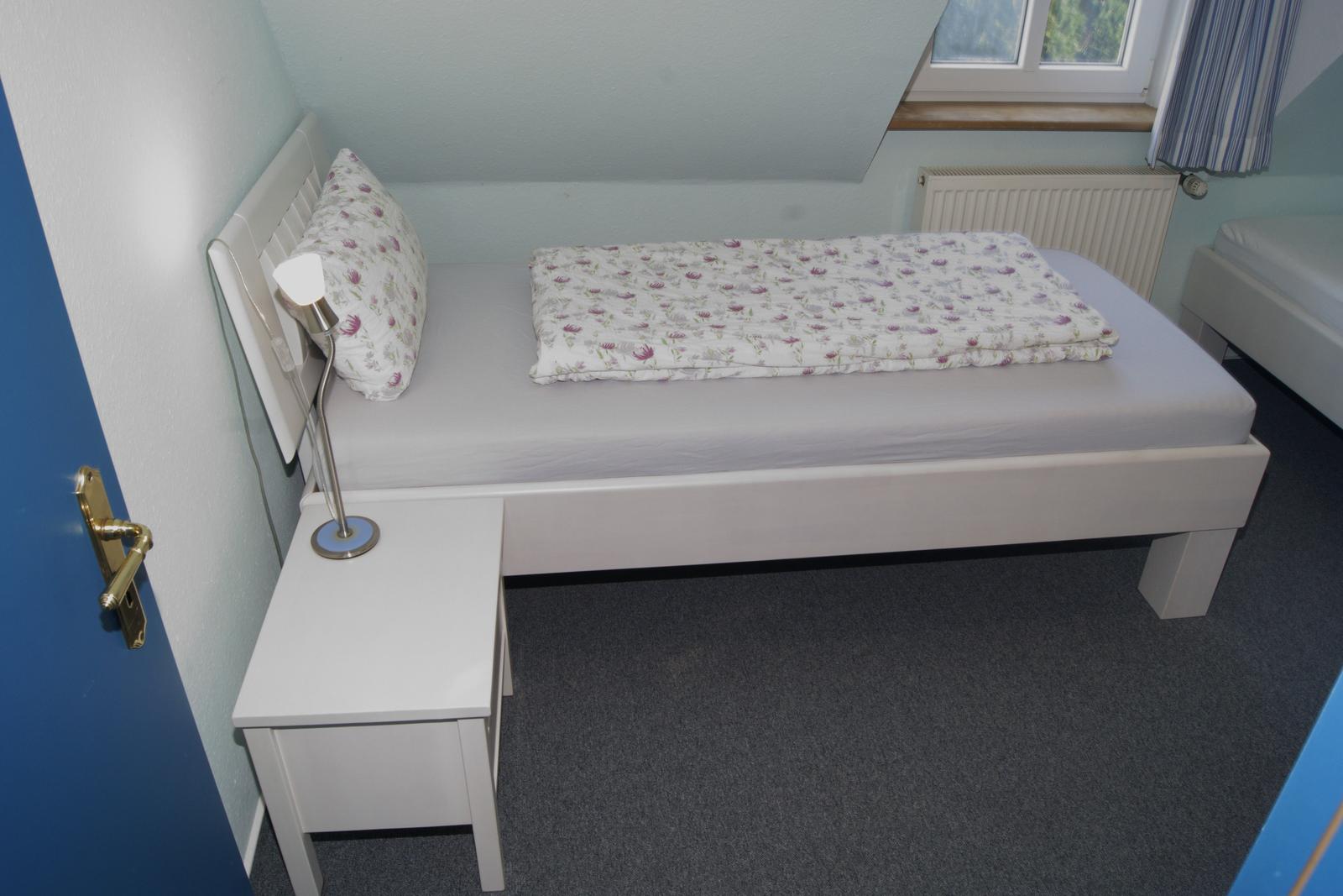Ein Schlafzimmer mit weißem Bett, Nachttisch und Lampe. Fenster und Heizung sind sichtbar.
