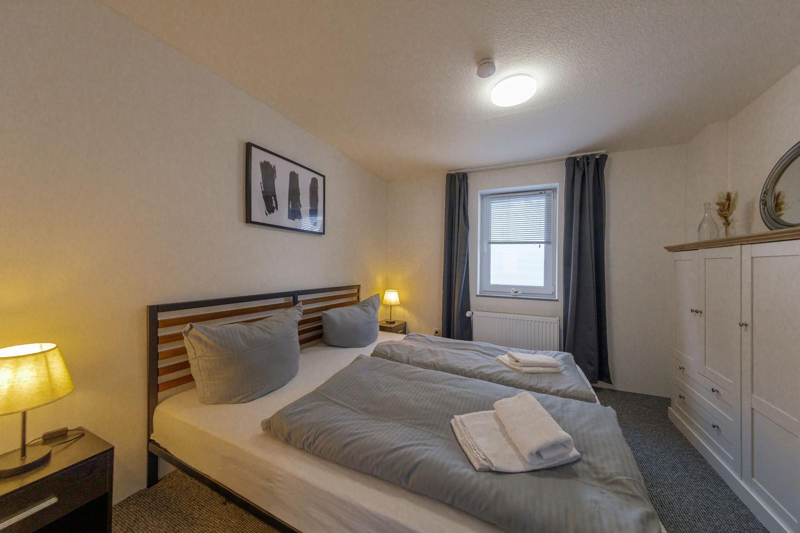 Schlafzimmer mit Doppelbett, Nachttisch, Kleiderschrank und Fenster mit Vorhängen.