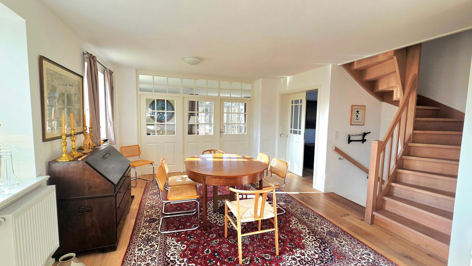 Esszimmer mit Holztisch, Stühlen und Teppich. Holzstufen führen nach oben.