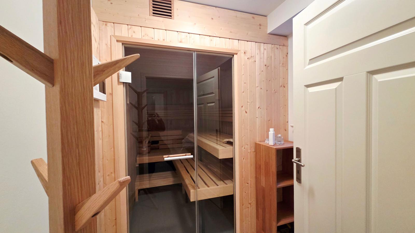 Sauna mit Holzwand, Glaswand und Holzbänken. Nebenraum mit Holzregal und Tür.
