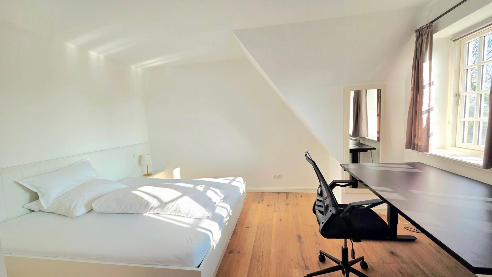 Schlafzimmer mit Dachschräge, Bett, Schreibtisch und Fenster.