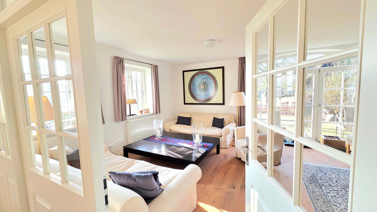 Wohnzimmer mit Sofa, Tisch und Fenster. Durchgangstür führt zum Garten.