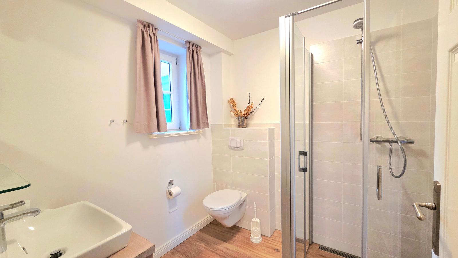 Badezimmer mit Dusche, WC und Waschbecken. Fenster mit Vorhängen.