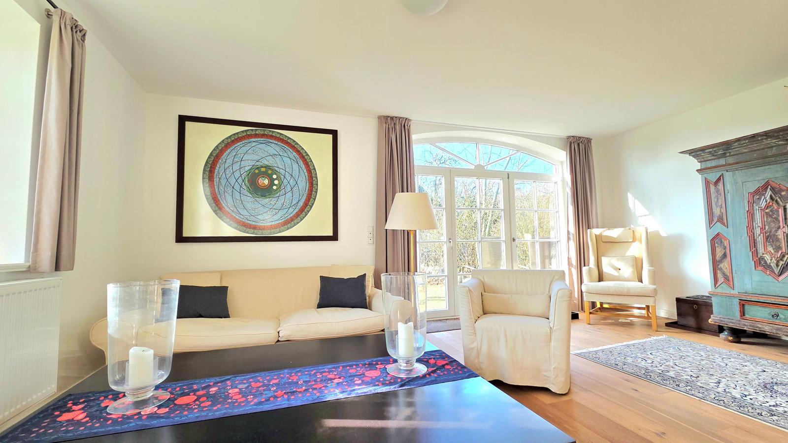 Wohnzimmer mit Sofa, Sessel und großem Fenster. Wandbild und antiker Schrank sind sichtbar.