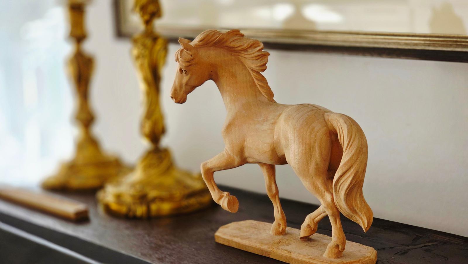 Holzfigur eines laufenden Pferdes auf dunklem Möbelstück mit goldenen Dekorationsobjekten im Hintergrund.