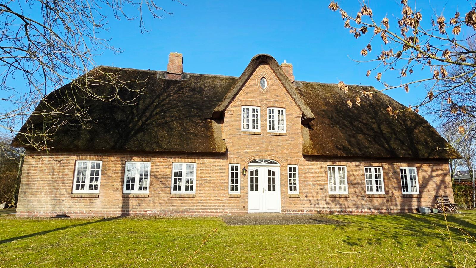Haus mit strohgedecktem Dach und rotem Backsteinbau. Weiße Fenster und Tür. Grasfläche vor dem Haus.