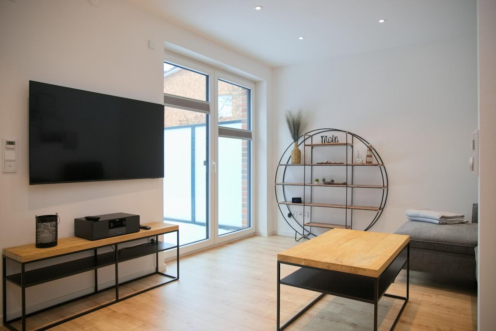 Wohnzimmer mit Fernseher, Tisch und Schrank. Große Fensterfront.