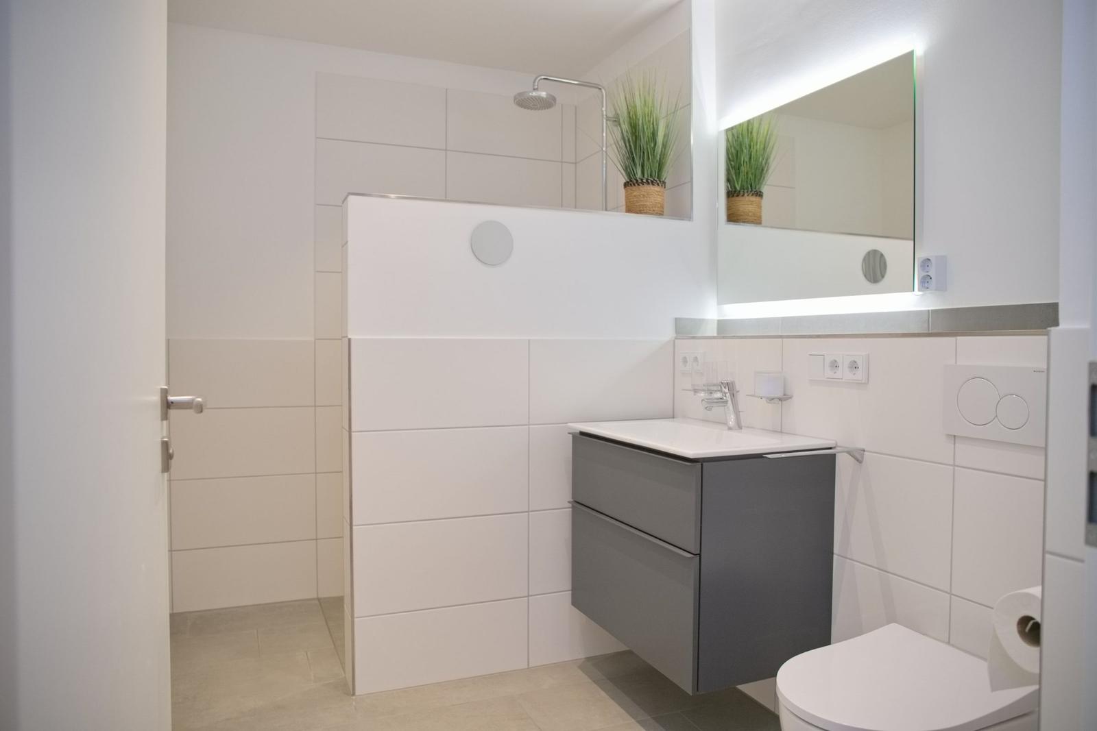 Badezimmer mit Dusche, Waschbecken und Toilette. Helle Fliesen und Spiegel mit Beleuchtung.