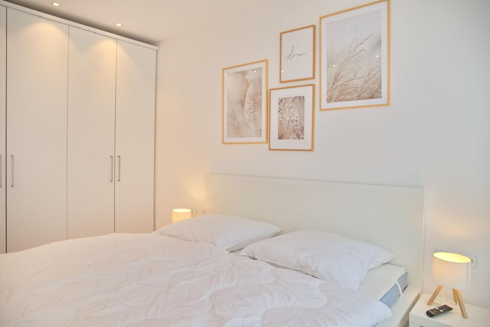 Schlafzimmer mit Bett, Nachttisch und Kleiderschrank. Wandbilder und Lampen sind angebracht.