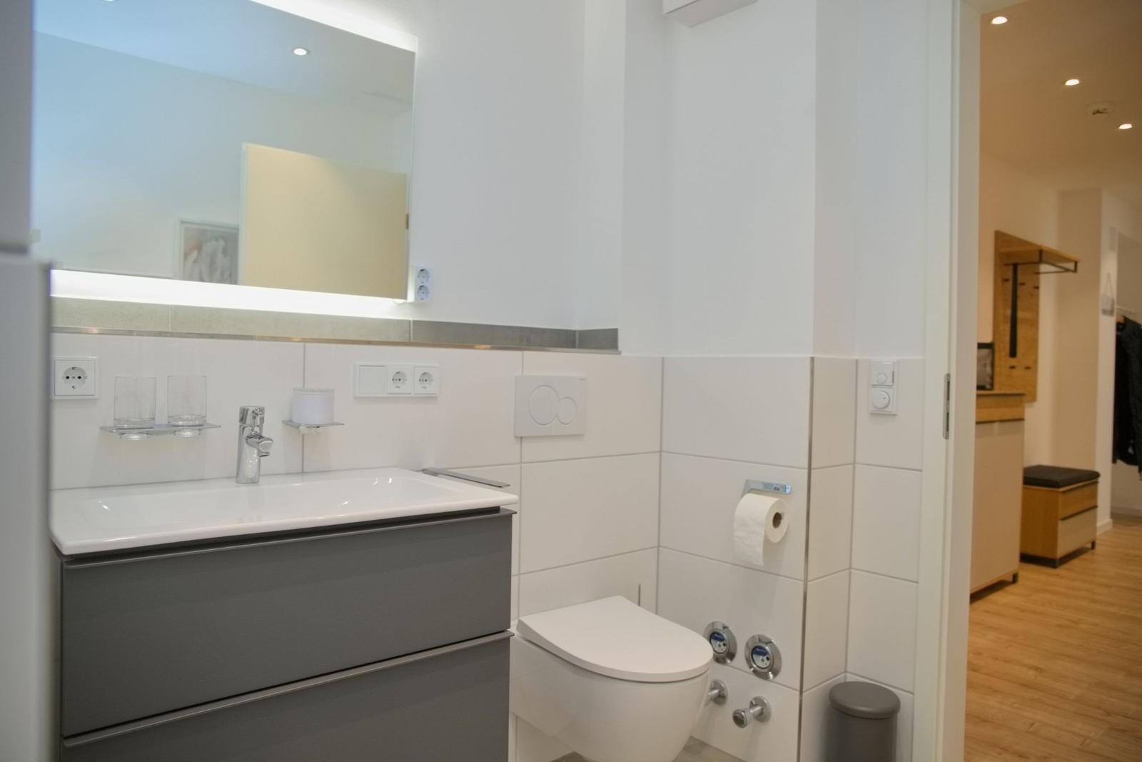 Badezimmer mit Waschbecken, Spiegel und Toilette. Blick in angrenzenden Raum.