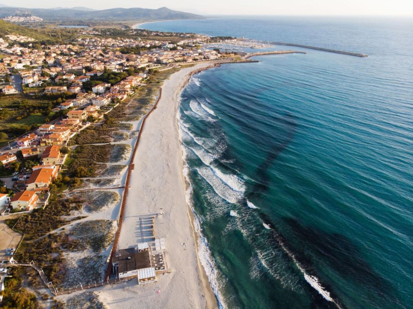 Strand mit Sand und türkisfarbenem Meer, Küstenstadt mit Häusern und Hafen im Hintergrund.