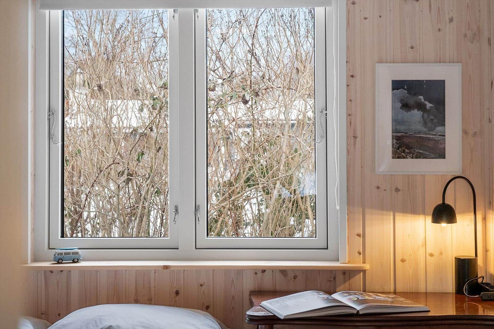 A window with a view of bare branches, beside a lamp and an open book.