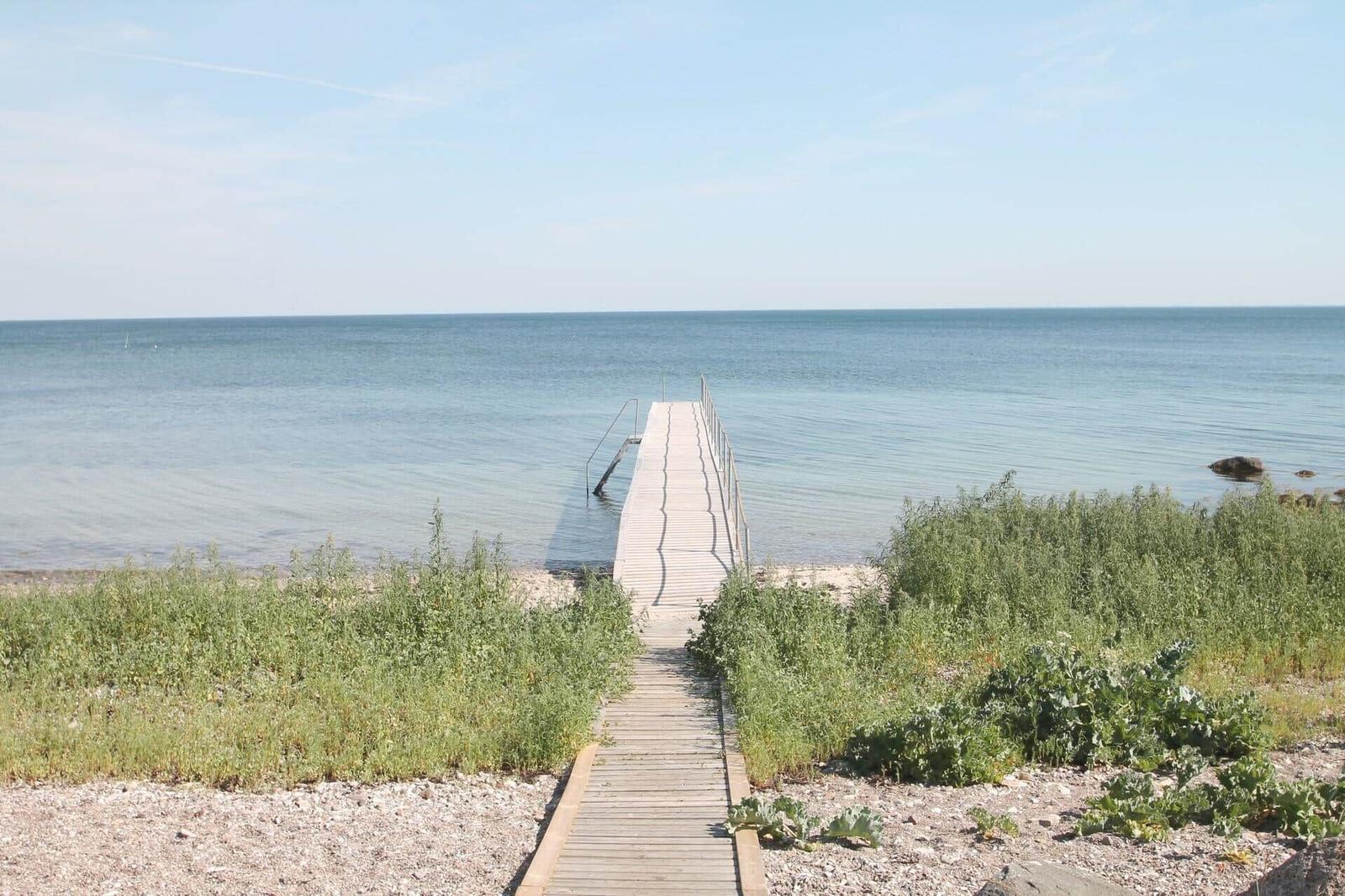 Wooden pier leads to water. Beach with grass and stones.