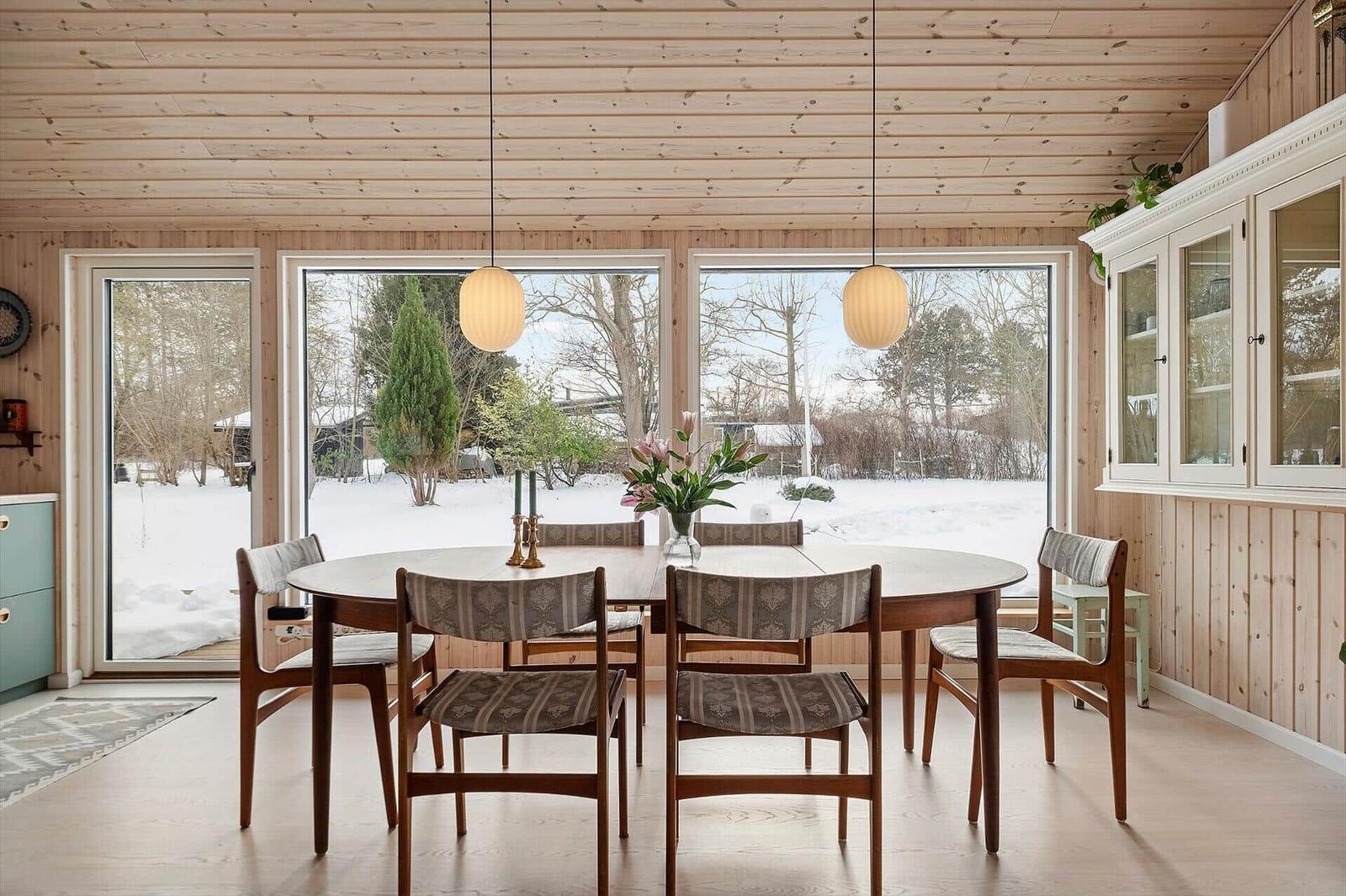 Dining room with round table, chairs, and large windows. Wooden ceiling and white cabinets.