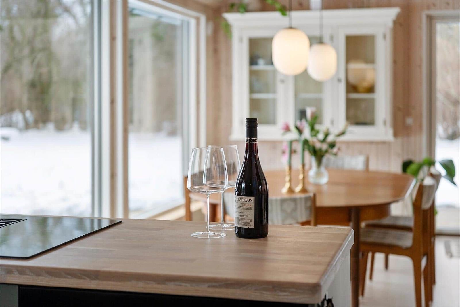 A table with wine bottle and glasses in dining area with wooden wall and window.