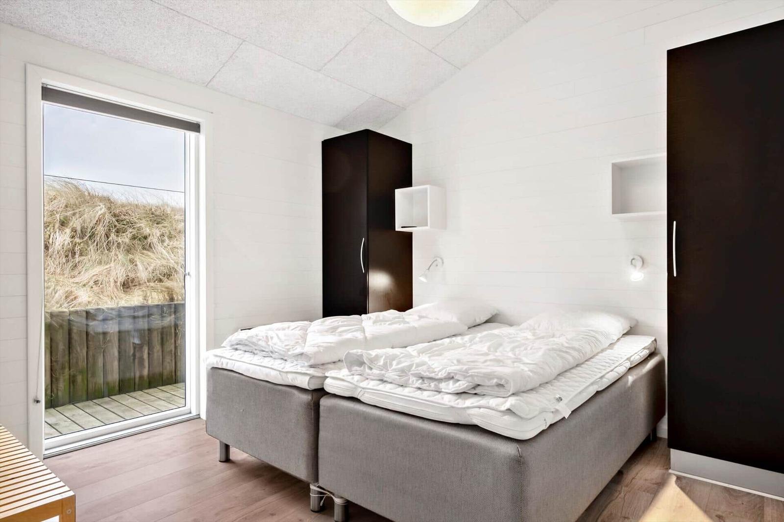 Schlafzimmer mit Doppelbett, dunklen Schränken und Blick auf Dünensand.